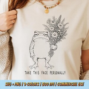 May include: A cream-colored t-shirt featuring a black line art illustration of a grumpy toad holding a large bouquet of wildflowers. Below the toad, the text reads "TAKE THIS FACE PERSONALLY" in a simple, bold, black sans-serif font.