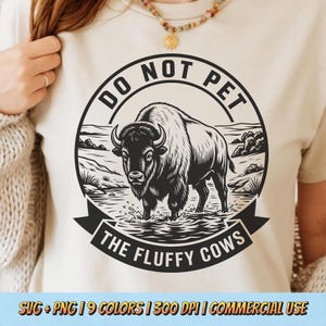 May include: Cream-colored t-shirt featuring a black and white illustration of a bison standing in water, surrounded by a landscape. The text "DO NOT PET" is arched above the bison, and "THE FLUFFY COWS" is below. The shirt also includes the text "SVG + PNG | 9 COLORS | 300 DPI | COMMERCIAL USE".