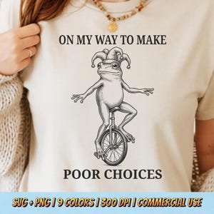 May include: Cream-colored t-shirt featuring a whimsical illustration of a frog riding a unicycle, wearing a jester's hat. The text "ON MY WAY TO MAKE POOR CHOICES" is printed above and below the frog. The shirt is a light neutral color.