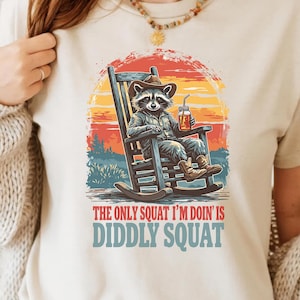 May include: A white t-shirt with a vintage-style illustration of a raccoon sitting in a rocking chair, holding a glass of iced tea. The text "The only squat I'm doin' is diddly squat" is printed below the illustration.