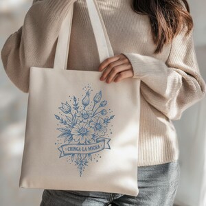 May include: Cream-colored tote bag with a blue floral design and the text "CHINGA LA MIGRA" on a banner. The design features various flowers and leaves. The bag has two long straps for carrying over the shoulder.