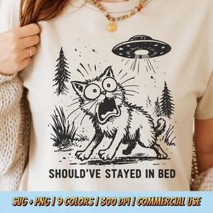Könnte beinhalten: Beigefarbenes T-Shirt mit einer schwarz-weißen Illustration einer erschrockenen Katze und einem UFO. Der Text "SHOULD'VE STAYED IN BED" ist darunter gedruckt. Das Shirt enthält auch den Text "SVG + PNG | 9 COLORS | 300 DPI | COMMERCIAL USE".