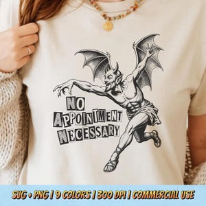 May include: A cream-colored t-shirt featuring a black graphic of a winged, horned figure in a dynamic pose. The text "NO APPOINTMENT NECESSARY" is printed in a bold, blocky font below the figure. The design has a vintage, etched illustration style.