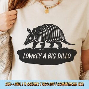 May include: A beige t-shirt featuring a black and white armadillo graphic. The armadillo is standing on a black oval with the text "LOWKEY A BIG DILLO". The shirt is a neutral color and suitable for casual wear.