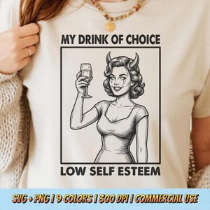May include: A beige t-shirt featuring a black and white graphic of a woman with devil horns holding a glass of wine. The text "MY DRINK OF CHOICE" and "LOW SELF ESTEEM" are above and below the image. The shirt is being worn by a person.
