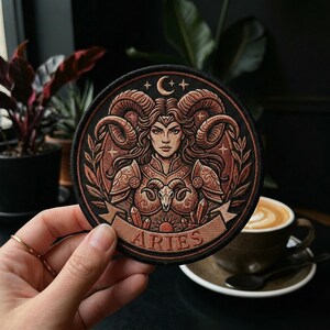 May include: A round, embroidered patch featuring the Aries zodiac sign. The design includes a woman with ram horns, a crescent moon, and the word "ARIES" on a banner. The patch is primarily brown and tan with black edging.