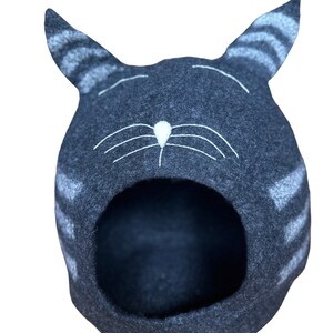 May include: A gray felt cat-shaped pet bed with white embroidered details. The bed has a large opening for a pet to enter.