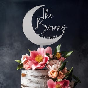 May include: White acrylic cake topper shaped like a crescent moon with the text "The Browns 12.10.2024" on it.