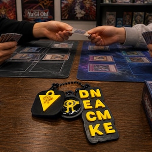 May include: A tabletop scene featuring a card game with players holding cards. A black and yellow keychain with the text "DECK" and other symbols is in the foreground. Card game mats and a card box are also visible.