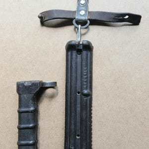 Op de afbeelding: Een donkerbruine, getextureerde bajonetschede van kunststof met een metalen bevestigingslus en een bruine leren riemhanger. Daarnaast ligt een bijpassend, donkerbruin bajonetvat van kunststof met een ronde metalen ring en een greep.