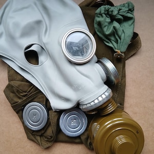 Op de afbeelding: Een grijze rubberen gasmasker met ronde oogstukken en een schroefdraadbus. Het masker wordt vergezeld door een groene canvas tas en twee metalen filterdoppen. Het masker is ontworpen om het hele hoofd en gezicht te bedekken.