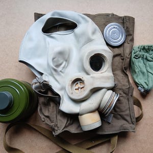 Op de afbeelding: Een vintage gasmasker met een lichtgrijze rubberen gelaatsmasker, ronde ooglenzen en een groene filterbus. Het masker wordt vergezeld door een canvas tas, een metalen deksel en een kleine groene pouch.