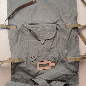 May include: Olive green canvas backpack with multiple straps and a front pocket. The bag features a leather tag and a flap pocket with faded white markings. The straps are a lighter shade of green.