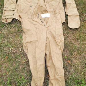 May include: A tan long-sleeved jacket and matching trousers set. The jacket features a button-up front, collar, and chest pockets. The trousers have a button fly, reinforced knee panels, and adjustable straps at the bottom of the pant legs.