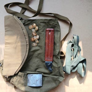 May include: An olive green canvas bag with a shoulder strap, a brown-handled knife, and several gold-colored star-shaped buttons. A silver-colored belt buckle with a star is also visible.