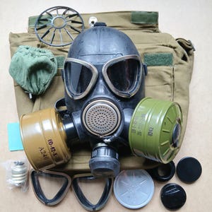 Puede incluir: Una máscara de gas negra con lentes grandes, dos filtros cilíndricos verdes y una bolsa de transporte de lona color canela. Componentes adicionales incluyen una cubierta de ventilación metálica, una bolsa de tela verde y varios accesorios negros y plateados.