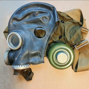 Puede incluir: Una máscara de gas negra con lentes transparentes y un filtro verde. La máscara va acompañada de una bolsa y correas de lona de color caqui. La máscara parece estar hecha de goma o un material similar.