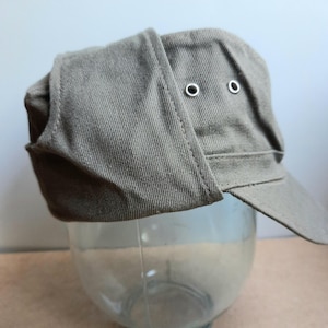 May include: A khaki-coloured cap with a peak and ear flaps. The cap has two metal eyelets on the top and is made of a textured fabric. The ear flaps are folded up. The cap is resting on a clear glass object.