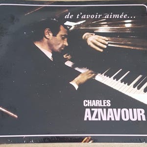 May include: Album cover featuring Charles Aznavour playing a piano. The image is in black and white, with the text "de t'avoir aimée..." at the top and "CHARLES AZNAVOUR" at the bottom. The cover has a vintage aesthetic.