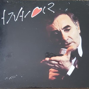 Peut inclure: Pochette d'album avec le nom "Aznavour" en écriture blanche et un cœur rouge. Un homme en costume sombre et chemise blanche tient un microphone sur fond noir.