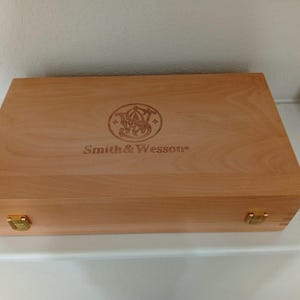 May include: A light-colored wooden box with the Smith & Wesson logo and text. The box has a rectangular shape with brass-colored metal clasps on the front. The wood grain is visible on the surface.