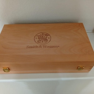 Può includere: Una scatola di legno color chiaro con il logo e il testo "Smith & Wesson" incisi sul coperchio. La scatola ha chiusure e cerniere in metallo color ottone. La venatura del legno è visibile.