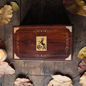 Pode incluir: Uma caixa retangular de madeira com acabamento castanho escuro, adornada com cantos dourados e acabamentos decorativos. A caixa apresenta o logotipo "COLT" com um cavalo empinado. Folhas de outono circundam a caixa.