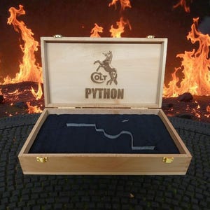 Könnte beinhalten: Geöffnete Holzkiste mit dem Colt Python Logo und Text. Die Kiste hat ein dunkelblaues Innenfutter mit einem Ausschnitt. Flammen sind im Hintergrund zu sehen.