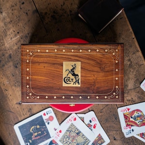Puede incluir: Una caja de madera con el logotipo de Colt y detalles dorados. La caja marrón de forma rectangular está sobre una superficie roja. Cartas de juego esparcidas sobre una mesa de madera.