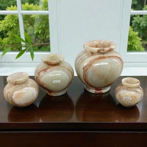 May include: Four decorative onyx vases of varying sizes sit on a dark wooden surface. The vases are a creamy white color with brown and orange veining. The smallest vase is a small, round shape, while the largest is a bulbous shape with a wide opening.