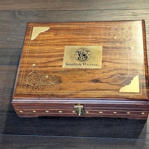 Pode incluir: Uma caixa retangular de madeira com acabamento em veios de madeira castanho-escuro. A tampa apresenta uma placa central de latão gravada com o logótipo e texto Smith & Wesson, detalhes dourados nos cantos e delicados arabescos dourados.