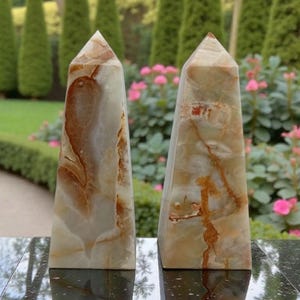 Könnte beinhalten: Zwei Onyx-Obelisken mit poliertem Finish. Die Obelisken haben eine hellbeige Grundfarbe mit braunen und beigen Adern. Die Obelisken werden auf einer reflektierenden schwarzen Oberfläche mit einem verschwommenen grünen und rosa Hintergrund präsentiert.
