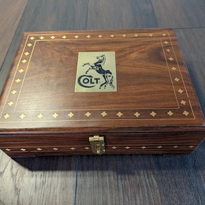 Peut inclure: Une boîte rectangulaire en bois brun foncé avec une plaque métallique dorée sur le couvercle, ornée d'un logo noir représentant un cheval cabré et du texte COLT. Le couvercle et le bord avant présentent une incrustation dorée en forme d'étoiles.