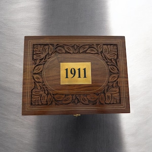 May include: A rectangular wooden box with intricate floral carvings around the edges. A gold-colored plaque in the center displays the number "1911". The box is made of dark brown wood with a polished finish.