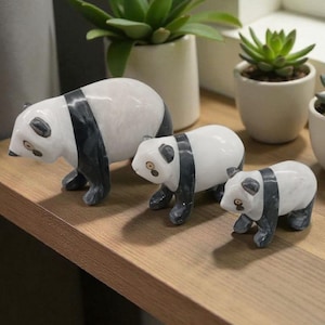 May include: Three panda bear figurines in varying sizes. The pandas are white with black markings and are posed in a walking position. The figurines are displayed on a wooden shelf, with small potted succulents in the background.