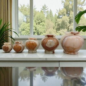 May include: Five decorative stone vases of different sizes, in shades of pink, brown, and cream, with unique veining. The vases are displayed on a white surface. The smallest vase is approximately 10 cm in height.