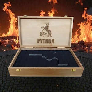 以下が含まれることがあります： Colt Pythonのロゴとテキストが入った木製の箱。箱は開いており、切り抜きのある濃い青色の内側が見えます。背景には炎があります。