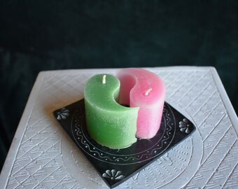 May include: A green and pink yin-yang shaped candle with white wicks, resting on a black square coaster with floral designs. The candle sits on a white, patterned surface, with a dark green background.