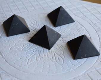 May include: Four black obsidian pyramids on a white surface with a decorative pattern. The pyramids are all the same size and shape.