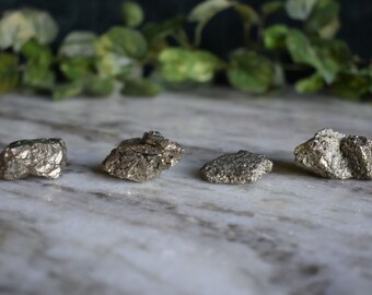 May include: Four raw pyrite crystals, also known as fool's gold, on a white marble surface. The crystals are various sizes and shapes, with a metallic silver color.