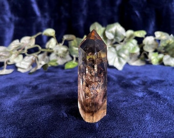 May include: A clear quartz crystal point with inclusions of brown and gold. The crystal is polished and has a pointed tip. It is sitting on a blue velvet surface.