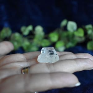 Puede incluir: Un cristal transparente y facetado con una textura rugosa y natural. El cristal est&aacute; descansando sobre una mano, con un anillo en el dedo.