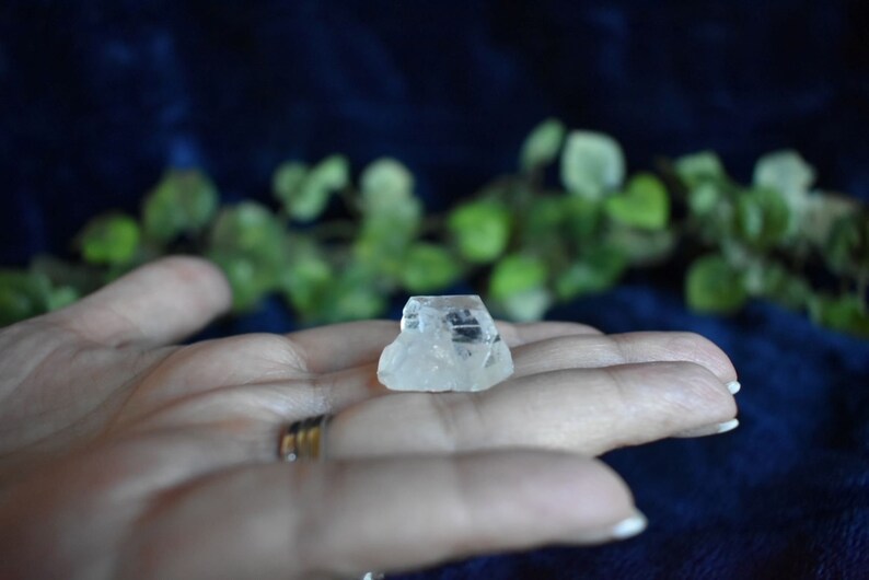 Puede incluir: Un cristal transparente y facetado con una textura rugosa y natural. El cristal est&aacute; descansando sobre una mano, con un anillo en el dedo.