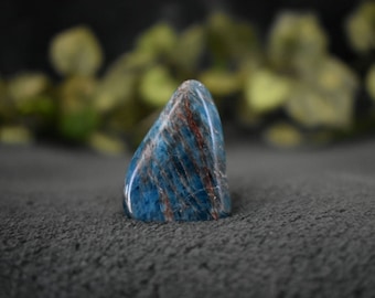 May include: A blue and brown polished gemstone with a smooth, rounded shape. The gemstone is sitting on a dark gray surface.
