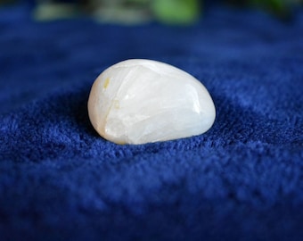May include: A smooth, white, oval-shaped crystal with a translucent appearance. The crystal is resting on a blue, fuzzy surface.
