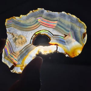 May include: A translucent agate slice with layered bands of white, grey, and brown, displaying iridescent rainbow hues. The natural stone features a dark, irregular hole and is held by a finger, showcasing its unique patterns and textures.
