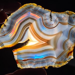 May include: A large, iridescent agate slice featuring a range of colours, including orange, brown, blue, and white. The agate displays a distinctive, swirling pattern and is backlit, showcasing its translucent quality.