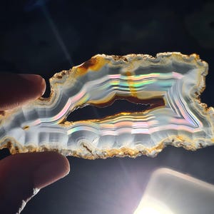 May include: A slice of agate with iridescent bands of colour. The agate is translucent and has a natural, rough edge.