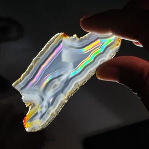 May include: A slice of iridescent agate displaying a rainbow of colours, including blue, green, and purple. The agate is translucent and has a white background.
