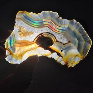 May include: A translucent agate slice, irregularly shaped, displaying bands of white, yellow, brown, and iridescent colours. The stone is held against a dark background, highlighting its natural patterns and textures.